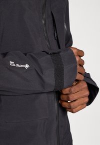 Casaco preto com fecho éclair na frente, apresentando uma faixa de velcro na manga e um logótipo na manga. Textura suave, design ajustado.