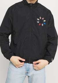 Svart zip-up jacka med elastiska manschetter, med färgglada lövmönster på vänstra bröstet. Tillverkad av lätt, texturerat tyg.