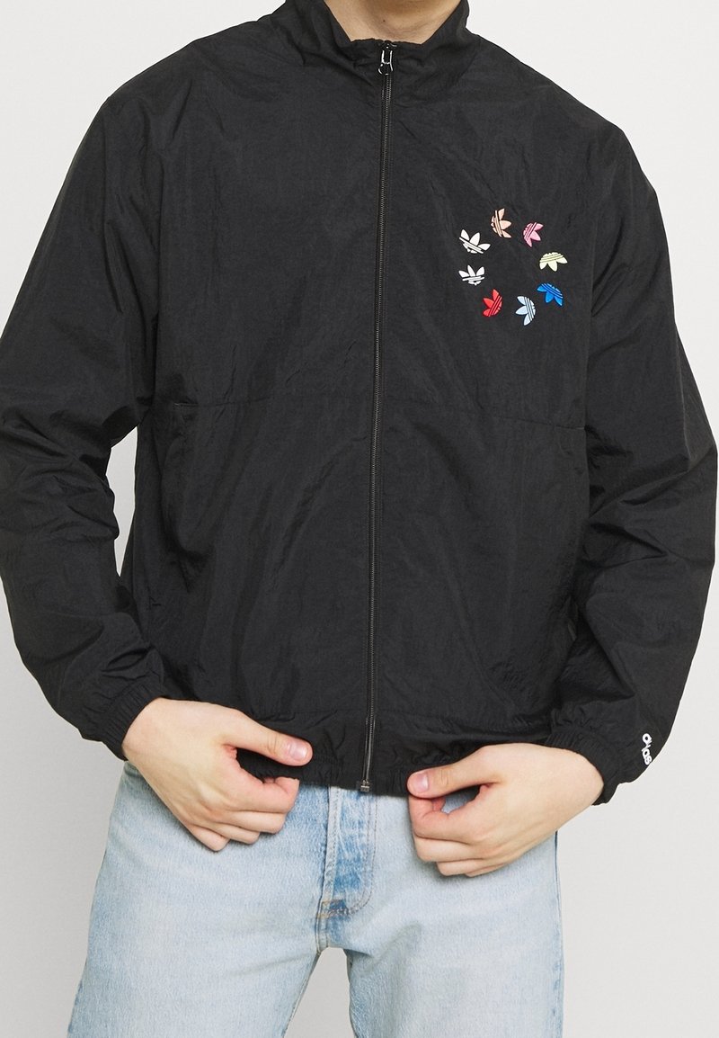 Svart zip-up jacka med elastiska manschetter, med färgglada lövmönster på vänstra bröstet. Tillverkad av lätt, texturerat tyg.