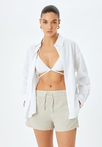 Hvid skjorte med knapper over en hvid bikini top og beige linned shorts. Skjorten har lange ærmer; shortsene har elastisk talje med snøre.