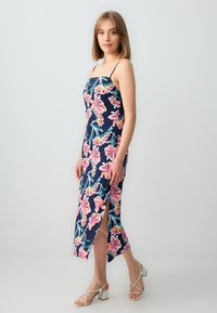 Junge Frau steht in einem floral gemusterten, navyblauen Midikleid mit seitlichem Schlitz und weißen Absatzsandalen vor einem schlichten Hintergrund.