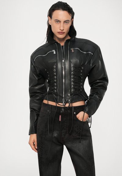 Sort læder cropped jakke med lynlås og snørebåndsdetaljer. Har høj krave og struktureret overflade. Stylet med mørke, mønstrede jeans.