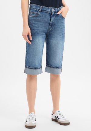 Osoba ubrana w niebieskie dżinsowe szorty o średniej długości z podwiniętymi nogawkami oraz białe trampki z czarnymi paskami, stojąca na jednolitym tle.