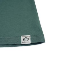 Grünes Baumwoll-T-Shirt mit flachem Bund. Ein gewebtes Etikett nahe dem Saum zeigt schwarze Schrift auf weißem Hintergrund und stellt überkreuzte Werkzeuge dar.
