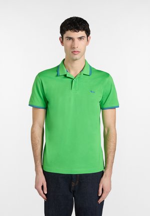 Giovane uomo in piedi, che indossa una polo verde brillante con rifiniture blu sul colletto e sulle maniche, abbinata a jeans blu scuro.