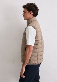 Leichte, beige gesteppte Weste mit hohem Kragen, die vertikale Steppabschnitte und eine Seitentasche mit Reißverschluss aufweist, über einem weißen Hemd getragen.