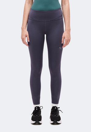 Ben iført mørkegrå tætsiddende leggings med Puma-logo nær venstre lår, sorte sportssko og hvide sokker på ensfarvet baggrund.