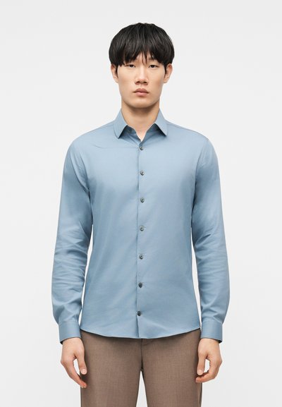 Man wearing a light blue long-sleeve button-up shirt and brown pants, facing forward with arms relaxed at sides.