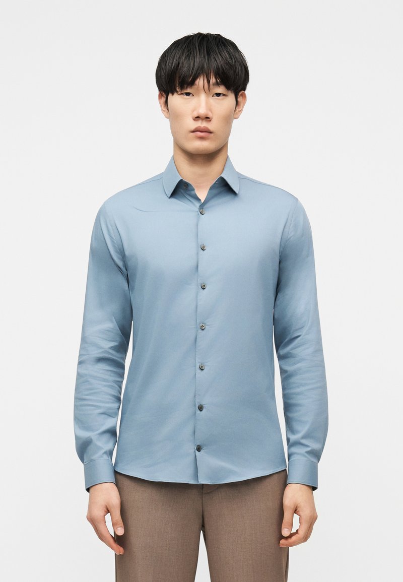 Άνδρας που φορά γαλάζιο πουκάμισο με μακριά μανίκια και καφέ παντελόνι, κοιτάζοντας μπροστά με τα χέρια χαλαρά στα πλάγια.