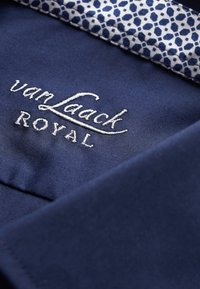 Navy fabric close-up featuring white embroidered "van Laack ROYAL" logo. Inner collar has a geometric navy and white pattern.