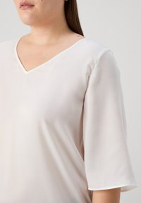 Lys beige bluse med V-hals, korte ærmer og glat tekstur. Tøjet ser blødt ud med minimal glans og subtile stikninger.
