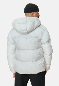 Doudoune blanche avec capuche, présentant un design matelassé et une texture gonflée. Le mannequin porte un bonnet noir et un pantalon foncé.