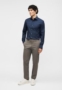 Άνδρας που στέκεται φορώντας μπλε πουκάμισο με κουμπιά, γκρίζο παντελόνι και λευκά αθλητικά παπούτσια, με τα χέρια στις τσέπες, κοιτάζοντας προς τα πλάγια.