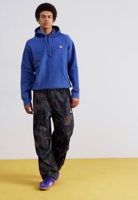 Moleton azul com cordão, calças com padrão de camuflagem e ténis roxos sobre um chão amarelo, modelo em pé contra um fundo simples.