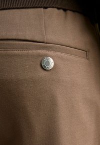 Gros plan d’un pantalon en tissu marron montrant une poche arrière horizontale avec un bouton rond argenté gravé "REISS".