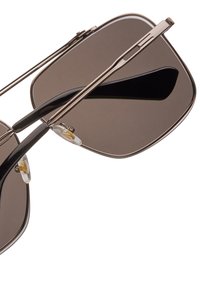 Sluneční brýle s tmavě hnědými čočkami, tenkým kovovým rámem, černými akcenty a průhlednými nosními podložkami. Unikátní skládací design s šikmými okraji.