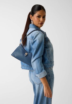 Vrouw in een spijkerjasje met studs en spijkerbroek draagt een kleine marineblauwe handtas over haar schouder en kijkt over haar schouder naar de zijkant.