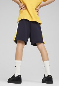 Marinblå shorts med gula och vita detaljer, kombinerade med svarta sneakers och vita ribbade strumpor. Tygkvaliteten ser mjuk ut och har en avslappnad passform.