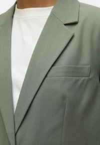 Blazer vert olive avec un revers cranté et une poche poitrine unique, fabriqué en tissu lisse, porté avec une chemise blanche à col rond en dessous.