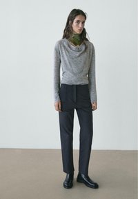 Maglione grigio a collo a cappuccio abbinato a pantaloni neri a vita alta e stivaletti neri alla caviglia. Il maglione ha una texture morbida; l'outfit è completato da una sciarpa verde a contrasto.