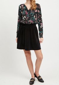 Blouse noire à motifs floraux avec manches longues, et jupe en velours noir avec une taille élastique. Mocassins noirs avec un accent de chaîne en or.
