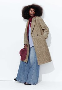 Manteau marron à motif chevrons, flares en denim bleu amples, superposé avec un col roulé bordeaux et une chemise bleue. Casquette de baseball bordeaux à la main.