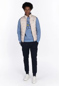 Joven con un chaleco acolchado beige sobre un sudadera azul con un gráfico de flores, pantalones cargo negros y zapatillas blancas, de pie con las manos en los bolsillos.