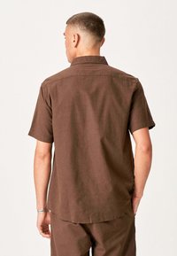 Mand iført en brun kortærmet skjorte med knapper og matchende bukser, der vender væk mod en ensfarvet lys baggrund.