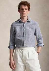 Άνδρας που φοράει μπλε και λευκό πουκάμισο με κάθετες ρίγες, με γυρισμένα μανίκια, λευκό πιετέ παντελόνι και ρολόι χειρός, στέκεται μπροστά σε απλό φόντο.