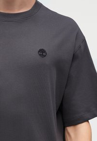 Σκούρο γκρι T-shirt με στρογγυλή λαιμόκοψη, με ένα μικρό μαύρο λογότυπο δέντρου κεντημένο στο αριστερό στήθος, φορεμένο από άτομο μπροστά σε απλό φόντο.