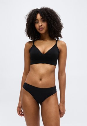 Γυναίκα με σγουρά μαλλιά που φοράει μαύρο σουτιέν και ταιριαστό μαύρο εσώρουχο, στέκεται μπροστά σε έναν απλό φωτεινό φόντο.