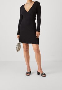 Mustapukuinen pitkähihaisia mekko, syvä V-kaula-aukko, sileä tekstuuri. Yhdistetty harmaisiin liukumekanavansandaaleihin, joissa on solki ja paksu korko.