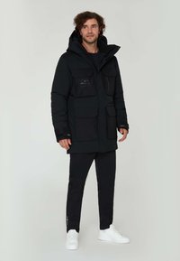 Svart isolerad parkas med huva, flera framfickor och dragkedja. Kombinerad med svarta byxor och vita sneakers. Slät tygtextur.