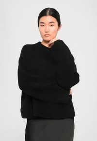 Giovane donna con capelli scuri raccolti indossa un maglione nero oversize e una gonna scura, in piedi con una mano vicino al mento su uno sfondo semplice.