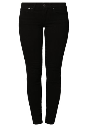 Osoba ubrana w dopasowany szary golf, czarne obcisłe spodnie i czarne buty na wysokim obcasie, stojąca z jedną ręką w kieszeni.