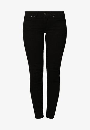 Persona vestida con un jersey de cuello alto gris ajustado, pantalones negros pitillo y zapatos de tacón negros, de pie con una mano en el bolsillo.