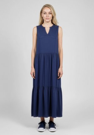 Kobieta w granatowej, bezrękawowej sukni maxi z marszczonymi warstwami oraz granatowych tenisówkach stoi na tle jasnoszarego tła.