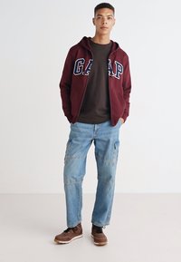 Sudadera burdeos con cremallera y logo "GAP", combinada con una camiseta marrón oscuro y vaqueros cargo azul claro. El modelo lleva zapatos marrones. Fondo simple.