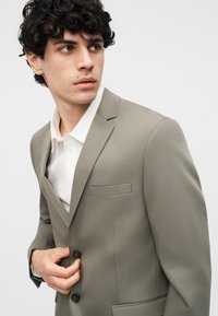 Νεαρός άνδρας με σγουρά σκούρα μαλλιά κουμπώνει τα κουμπιά σε ένα ανοιχτό γκρι σακάκι πάνω από ένα λευκό πουκάμισο, κοιτάζοντας προς τα αριστερά του.