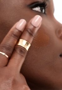 Närbild av en hand som applicerar bronzer på en slät, mörk hudton. Personen har två ringar på handen: en tunn silverring och en tjock guldring.