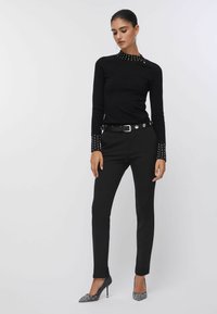 Haut noir à manches longues avec col et poignets ornés, associé à un pantalon noir ajusté et des talons pointus avec des accents scintillants.