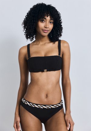 Kohaga naise lokkis juustega, kes kannab musta kaheosalist ujumistrikood, mille allosas on palmikutega valge ja beež muster, seisab ühevärvilise heleda tausta ees.