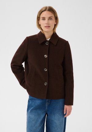 Femme portant un manteau marron foncé boutonné avec col et un jean bleu, debout avec une main derrière le dos sur un fond uni.