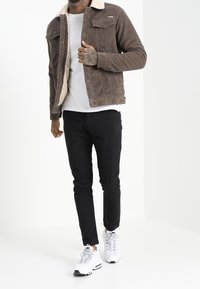 Ruskea corduroytakki, jossa on beige tekoturkiskaulus ja etutaskut, yhdistetty mustiin housuihin ja valkoisiin tennareihin, malli neutraalissa taustassa.