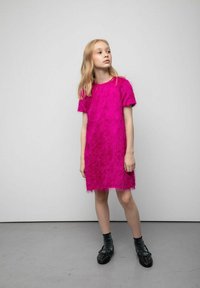 Fuksjowa sukienka o puszystej fakturze, z krótkimi rękawami i okrągłym dekoltem, zestawiona z czarnymi, ćwiekowanymi butami oraz wzorzystymi skarpetkami.