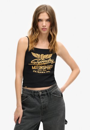 Zwarte geribbelde tanktop met gouden opdruk "Superdry 1st Class Motorsport" logo met vleugeldesign. Gecombineerd met hoogwaisted grijze denim jeans.