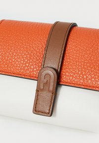 Orange texturerad läderpouch med vit bas, prydd med ett brunt band och en präglad logotyp på bandet.