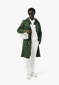 Groene trenchcoat over een witte hoodie en details, gecombineerd met witte broek en off-white sneakers. Accessoires omvatten een zonnebril en een schoudertas.