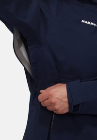 Navy waterproof jacket with a smooth texture, zippered side pocket, and Velcro cuff detail. Features a prominent brand logo in white.