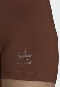 Hnědé látkové šortky s stříbrným rhinestonovým logem Adidas trefoil blízko lemu na levém stehně.